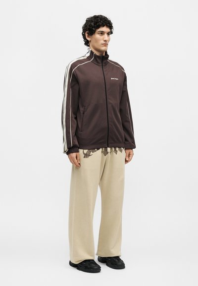 Άνδρας μοντέλο στέκεται, φορά καφέ ζακέτα με φερμουάρ και λευκές ρίγες και μπεζ φαρδιά παντελόνια με μαύρα αθλητικά παπούτσια.