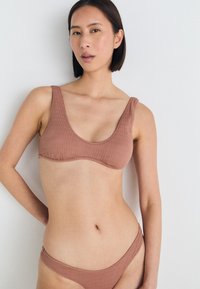 Ruda tekstūruota bikini maudymosi kostiumėlis su apvalia iškirpte ir plačiomis petnešėlėmis. Komplekte yra atitinkami žemo liemens bikini dugnai. Paprastas ir minimalistinis dizainas.