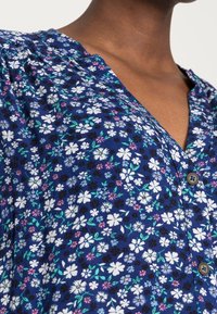 Bluse mit floralen Druck, die einen blauen Hintergrund und kleine weiße, rosa und grüne Blumen zeigt. Knopfleiste im V-Ausschnitt, weicher Stoff.