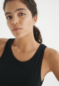 Musta tank top, jossa on pyöreä pääntie. Kangas näyttää sileältä ja kevyeltä, istuen tiiviisti vartalon ympärille ilman näkyviä kuvioita tai koristeita.