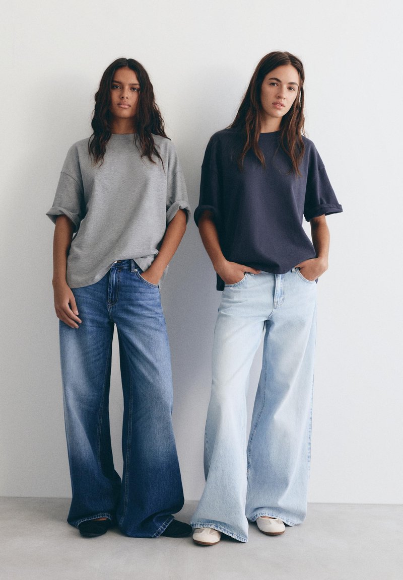 Dvije žene stoje jedna pokraj druge noseći traperice širokih nogavica, jedna u tamnoj boji s sivom majicom, druga u svijetloj boji s tamnoplavom majicom, ruke u džepovima.