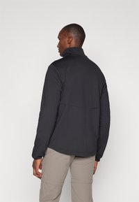 Giacca in pile nera con colletto alto, caratterizzata da una texture liscia e una cucitura sulla schiena. Indossata con pantaloni chiari.