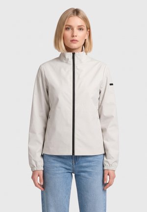 Donna che indossa una giacca grigia chiaro con zip, collo alto e polsini elasticizzati, abbinata a jeans azzurri, in piedi davanti a uno sfondo semplice.