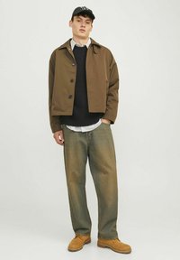 Braune kurze Jacke mit schwarzen Knöpfen, schwarzer strukturierter Pullover, gestreiftes Hemd, lockere grünliche Hose und beige Stiefel. Lässiger Streetwear-Stil.