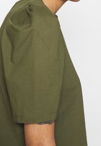 Oliven grøn t-shirt med korte ærmer, der har en rund halsudskæring og blødt, glat bomuldsstof. Sidevisningen viser skulderdetaljer med samling.