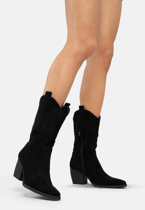Bottes noires en daim montantes, à bout pointu, talon empilé et coutures décoratives. Comprend une fermeture éclair sur le côté et des détails en boucle en haut.