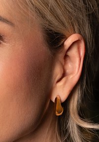 Goldfarbener Metalldraht mit einem abstrakten, geschwungenen Design. Die Oberfläche wirkt glatt und hat ein poliertes Finish. Nahaufnahme am Ohr.