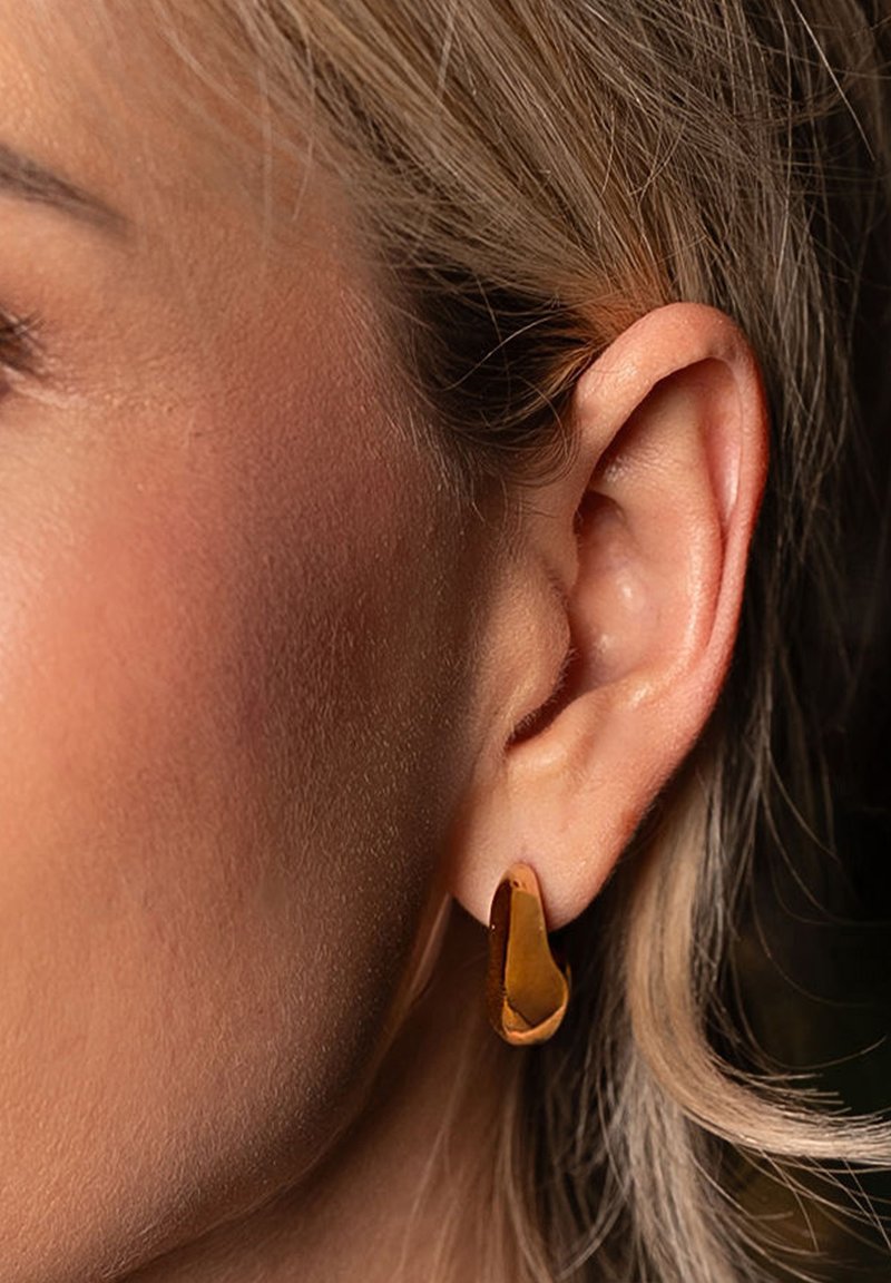 Pendiente de metal color dorado con un diseño abstracto y curvado. La superficie parece lisa, con un acabado pulido. Vista en primer plano en la oreja.