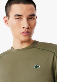 Tricou verde măsline dintr-un material neted, cu un guler rotund și un mic logo brodat cu un crocodil pe piept.