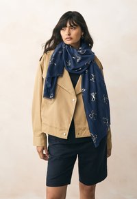 Sciarpa blu navy con motivi di fiocchi ricamati in oro, drappeggiata su una giacca beige e un top nero, indossata con pantaloncini blu navy. Tessuto morbido e dalla texture evidente.