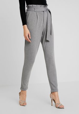 Vrouw draagt hoogwater grijze taps toelopende broek met een stoffen riem, gecombineerd met zilverkleurige sandalen met hoge hak en een zwarte geribbelde top.