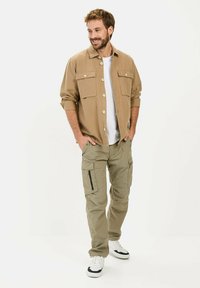 Tan overhemd met knopen en twee borstzakken, gecombineerd met lichtgekleurde kaki cargobroek met zijzakken en zwarte ritsen. Witte sneakers inbegrepen.