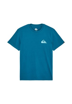 Man draagt een blauw T-shirt met korte mouwen met een klein wit logo op de linkerborst en beige broek, kijkt naar rechts.