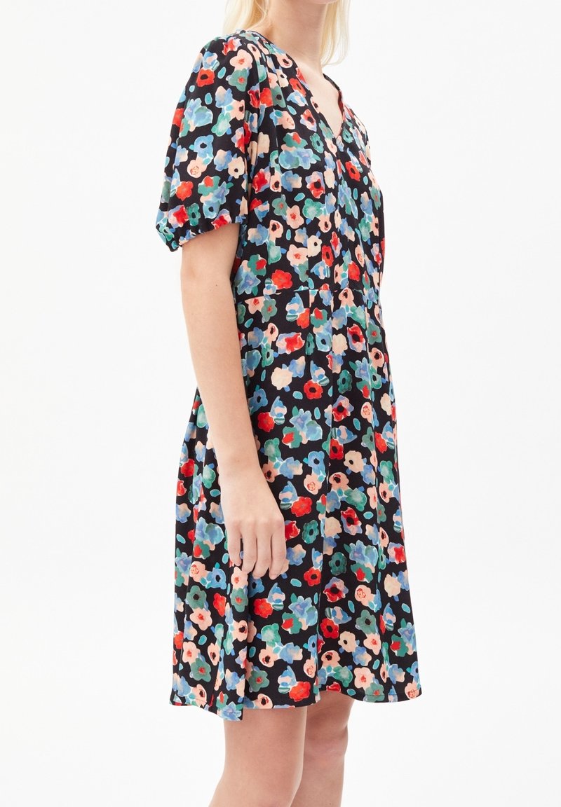 Femme portant une robe noire longueur genou avec imprimé floral rouge, bleu, vert et beige et manches courtes bouffantes sur fond blanc.