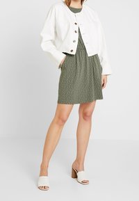 Veste blanche courte, robe verte à pois blancs, et mules blanches tissées à talons. Accent sur les boutons et les éléments de design décontractés.
