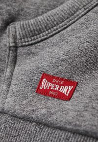 Tejido de sudadera gris con un acabado texturizado, que presenta un parche de logo bordado en rojo con la etiqueta "SUPERDRY 2003" en el cuello.