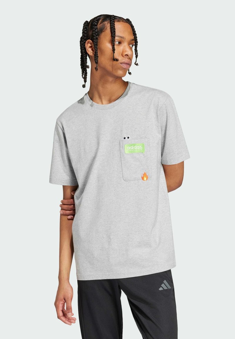 Mlad muškarac s pletenom kosom nosi svijetlosivu Adidas majicu s prednjim džepom, zelenim logotipom i malim grafičkim prikazom plamena, uparen s crnim hlačama.