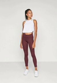 Biely skrátený top bez rukávov spárovaný s vínovými legínami, oboje z elastického materiálu. Modelka má na sebe biele tenisky a vlasy má upravené do vrkočov.