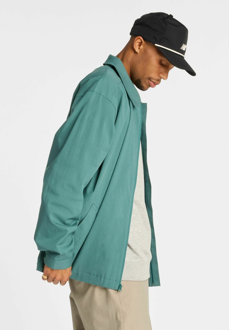 Leichte türkisfarbene Jacke mit lockerem Schnitt, ausgestattet mit einem spitzen Kragen, Seitentaschen und langen Ärmeln, kombiniert mit einer schwarzen Mütze.