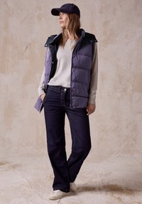Vadderad lila väst med marinblå detaljer över en ljusgrå tröja, kombinerad med mörka jeans och vita skor. En avslappnad keps fulländar looken.
