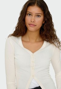 Cardigan a maniche lunghe color crema con coste, scollatura a V, cinque bottoni frontali e design aderente. Tessuto morbido e leggero.