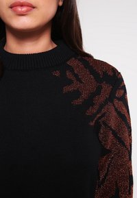 Pull noir avec un col côtelé et des accents de motif animalier métallique marron sur les manches, mettant en valeur un design texturé.