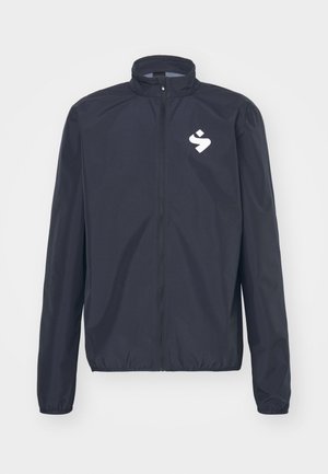 Veste coupe-vent navy avec un col haut, fermeture éclair complète, poignets élastiques et un logo blanc sur la poitrine gauche. Légère, texture lisse.