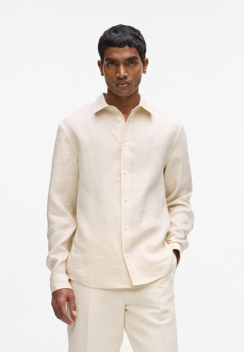 Chemise en lin beige clair avec col classique, manches longues et fermeture boutonnée sur le devant. Détail subtil de logo brodé près de la poitrine.