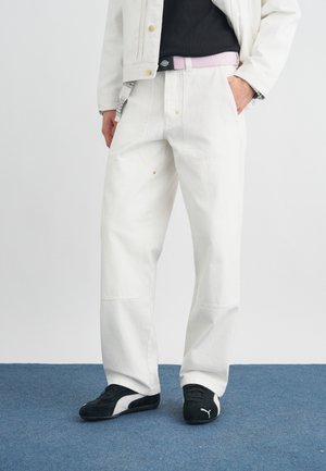 Osoba oblečená v bielych nohaviciach so širokými nohavicami, čierno-bielych teniskách a bielej bunde, s jednou rukou v vrecku na modrom koberci.