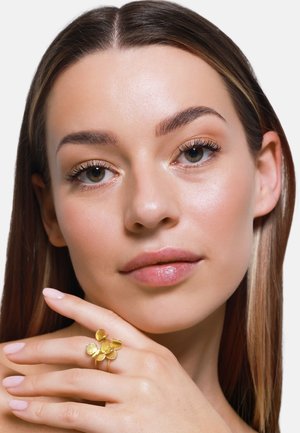 Goldener Blumenring mit fünf blütenblattförmigen Akzenten, getragen an einer Hand mit hellrosa Nägeln, vor einem neutralen Hintergrund. Glatte Hauttextur.