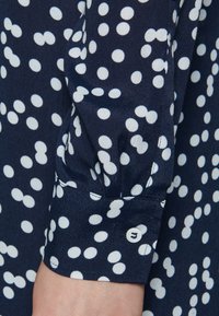 Blusa blu navy con pois bianchi, con polsini dettagliati da un bottone e un tessuto morbido e testurizzato. Inquadratura ravvicinata della zona delle maniche.