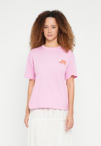 Tricou roz deschis din bumbac, cu mâneci scurte, decolteu rotund și un mic logo portocaliu pe piept. Design simplu, croială casual.