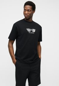 Νεαρός άνδρας που φοράει μαύρο σορτς και μαύρο μπλουζάκι με γραφικό λευκών γυαλιών ηλίου με σχέδιο και κείμενο "KARL LAGERFELD".