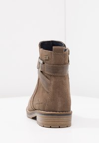 Bottine en daim marron avec un bout arrondi, une languette de tirage à l'arrière, un accent de sangle en cuir foncé et une semelle en caoutchouc avec des rainures horizontales.