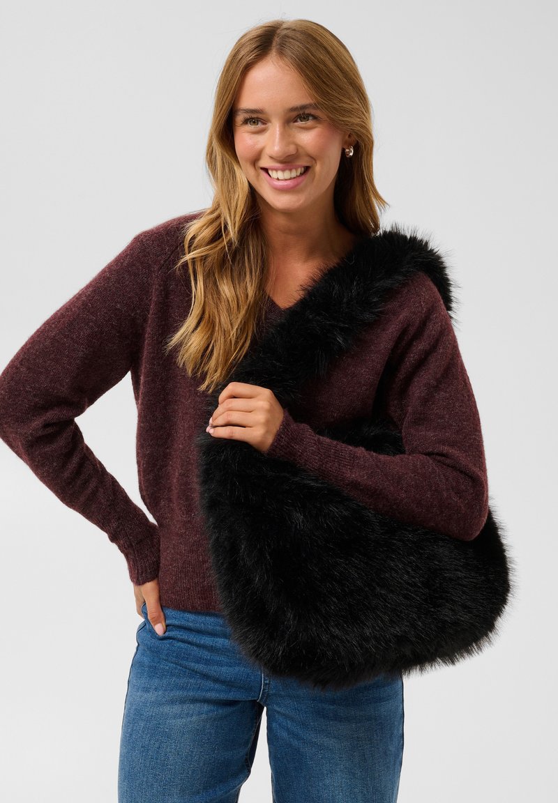 Jeune femme souriante portant un pull violet et un jean, tenant un grand sac à main noir et duveteux sur son épaule.