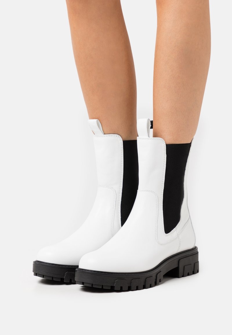 Botas de tobillo de cuero blanco con paneles laterales elásticos negros, punteras redondeadas y suelas de goma negra gruesas con un patrón de relieve.