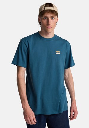 Jonge man met lichtbruin haar draagt een beige pet achterstevoren, een blauw T-shirt met korte mouwen en een klein oranje-crèmekleurig golflogo, en donkere broek.