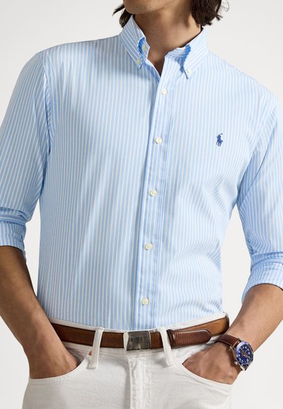 Ανοιχτό μπλε ριγέ πουκάμισο με λευκές ρίγες, γιακά με δύο κουμπιά και μικρό κεντημένο λογότυπο. Συνδυάζεται με σκούρα καφέ ζώνη και ανοιχτόχρωμο παντελόνι.