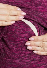 Des mains aux ongles vernis en rose ajustant l'ouverture d'une robe de maternité violette pour révéler un soutien-gorge d'allaitement en dessous.