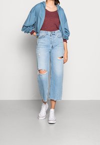 Ljusblå vida jeans med slitna knän, i kombination med en vinröd topp och en lös blå zip-up hoodie, avslutat med vita sneakers.