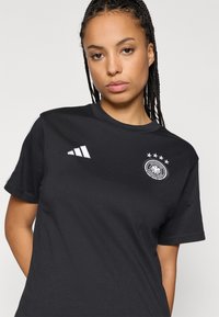 Zsinórra font hajjal rendelkező nő, fekete Adidas pólót visel, amelyen a Német Labdarúgó Szövetség logója és négy csillag látható a mellkason.