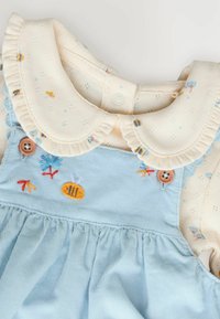 Vestido de pana azul claro con un cuello bordado en crema. Presenta bordes con volantes, patrones florales y botones de madera.