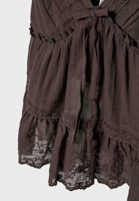 Vêtement en tissu marron avec bordure en dentelle florale brodée et un détail de nœud noué à la taille, mettant en valeur la texture et un design en couches.