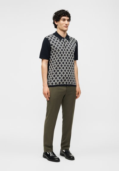 Άνδρας που φοράει μαύρο πόλο με κοντά μανίκια και άσπρο γεωμετρικό σχέδιο, χακί παντελόνι και μαύρα επίσημα παπούτσια, στέκεται μπροστά σε λευκό φόντο.