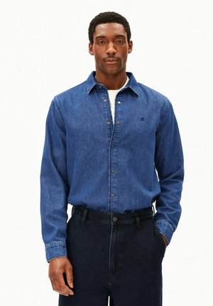 Mann trägt ein blaues Jeanshemd mit Knöpfen über einem weißen Unterhemd und dunklen Jeans, steht mit einer Hand in der Tasche vor einem einfarbigen Hintergrund.