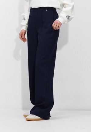 Persoon draagt losse marineblauwe broek, wit langarmshirt met bloemrijke borduursels en witte sneakers met gumzolen.