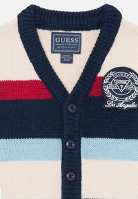 Navy ja vaalean kerman raidallinen neulecardigan, jossa on punaisia ja vaaleansinisiä kaistaleita. Tässä on V-pääntie, viisi nappia ja brodeerattu logopatch.
