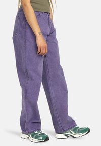 Pantaloni in denim viola con una vestibilità relaxed, caratterizzati da tasche laterali e dettagli di cucitura in vista. Indossati con sneaker verdi.