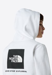 Hanorac alb cu un grafic pătrat negru pe spate, care prezintă logo-ul „The North Face” și textul „NEVER STOP EXPLORING” în alb.