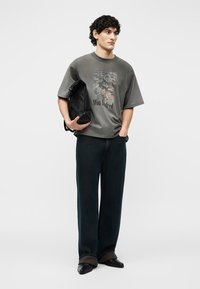 Młody mężczyzna stoi, trzymając czarną złożoną torbę, ubrany w dużą szarą koszulkę z nadrukiem, luźne ciemne dżinsy i czarne buty na białym tle.